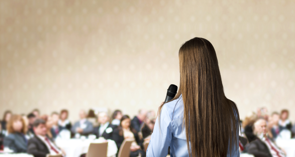 Woman with long brown hair and blue shirt presenting in front of professional audience.